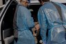 Two health care workers wearing blue protective gowns and face shields lean into an open vehicle door to examine a seated woman holding a baby.