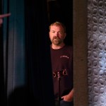 Graham Platner stands partly hidden behind a stage curtain in a dark venue as he looks toward the camera.