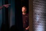 Graham Platner stands partly hidden behind a stage curtain in a dark venue as he looks toward the camera.