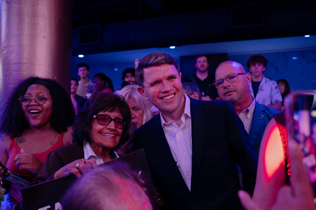 James Talarico in a dark suit poses for a photo with supporters in a crowded, dimly lit venue washed in blue and purple light.