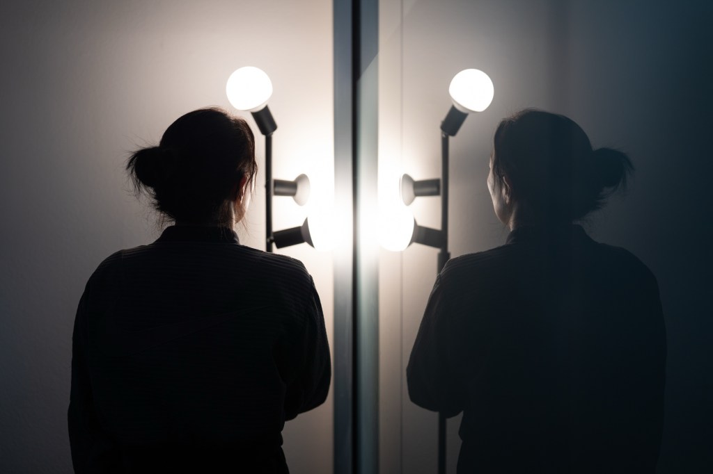 An anonymized person is reflected in a mirror image of them facing a lamp in silhouette.