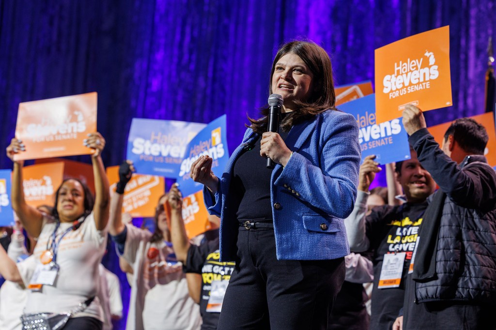 Senate candidate Haley Stevens speaks into a microphone onstage as supporters behind her hold orange and blue campaign signs.