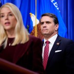 Pam Bondi speaks at a Justice Department podium in the foreground as Todd Blanche stands behind her, framed by American and departmental flags.