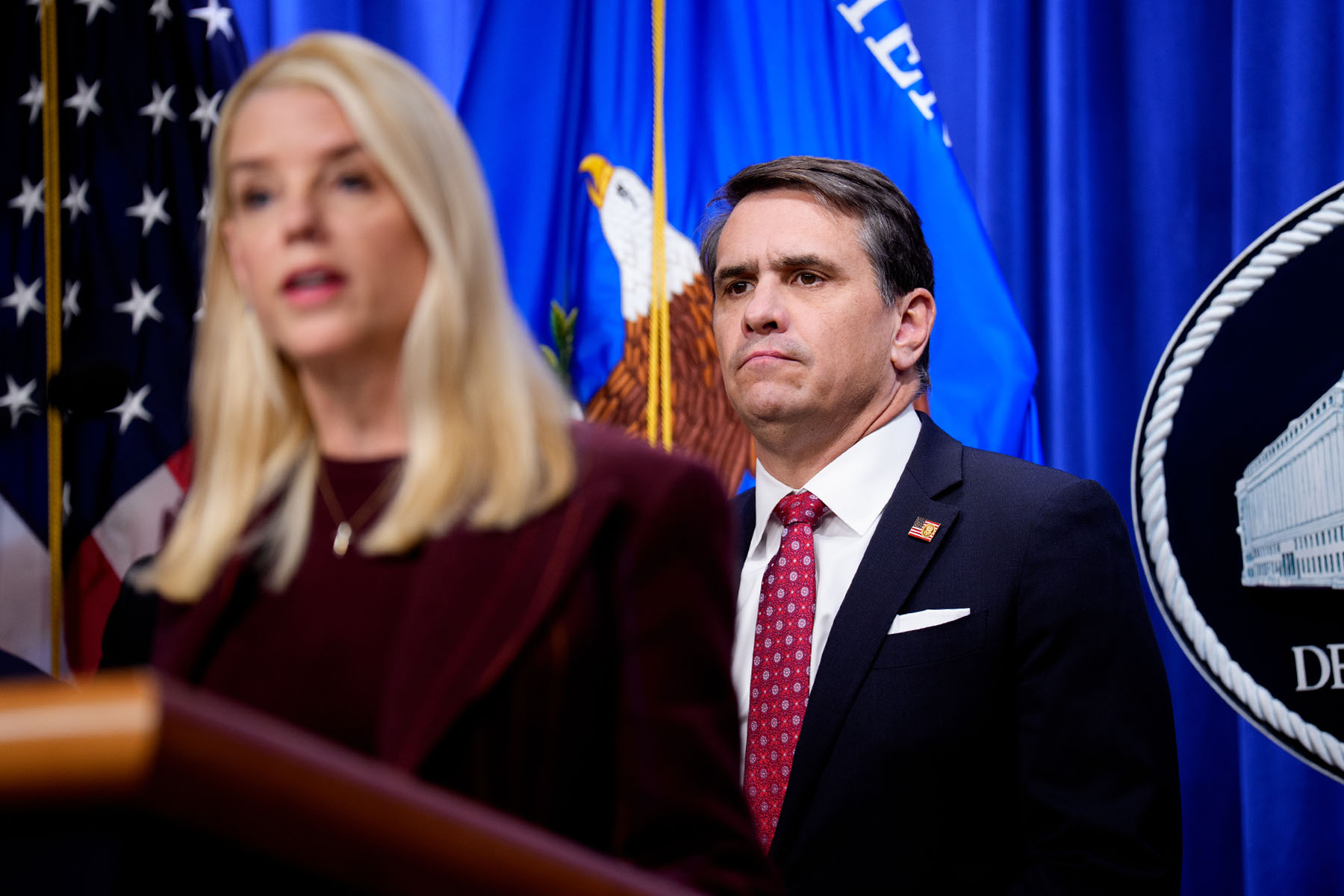 Pam Bondi speaks at a Justice Department podium in the foreground as Todd Blanche stands behind her, framed by American and departmental flags.