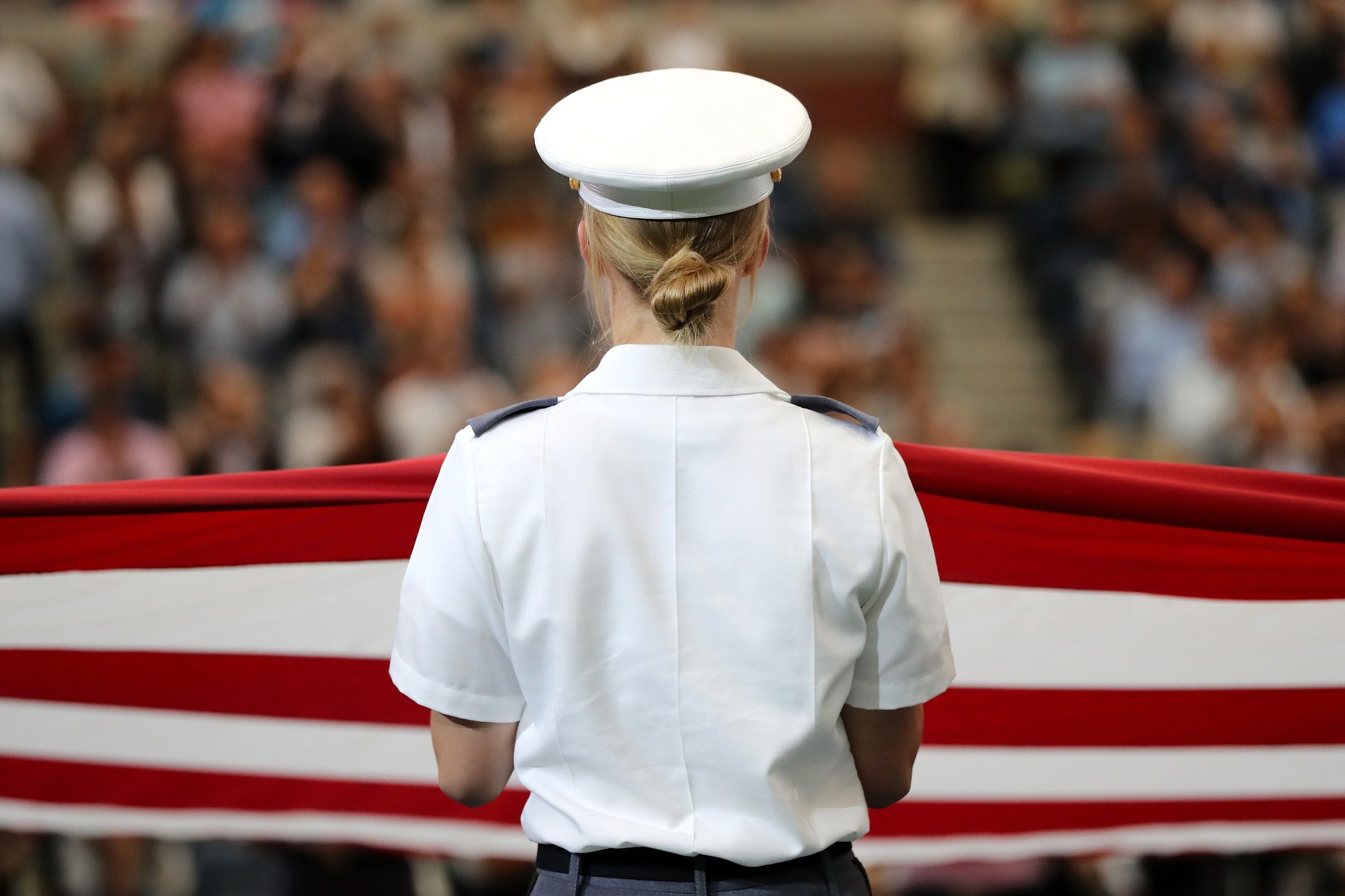 A woman in military uniform is seen from behind facing an audience and the US flag.
