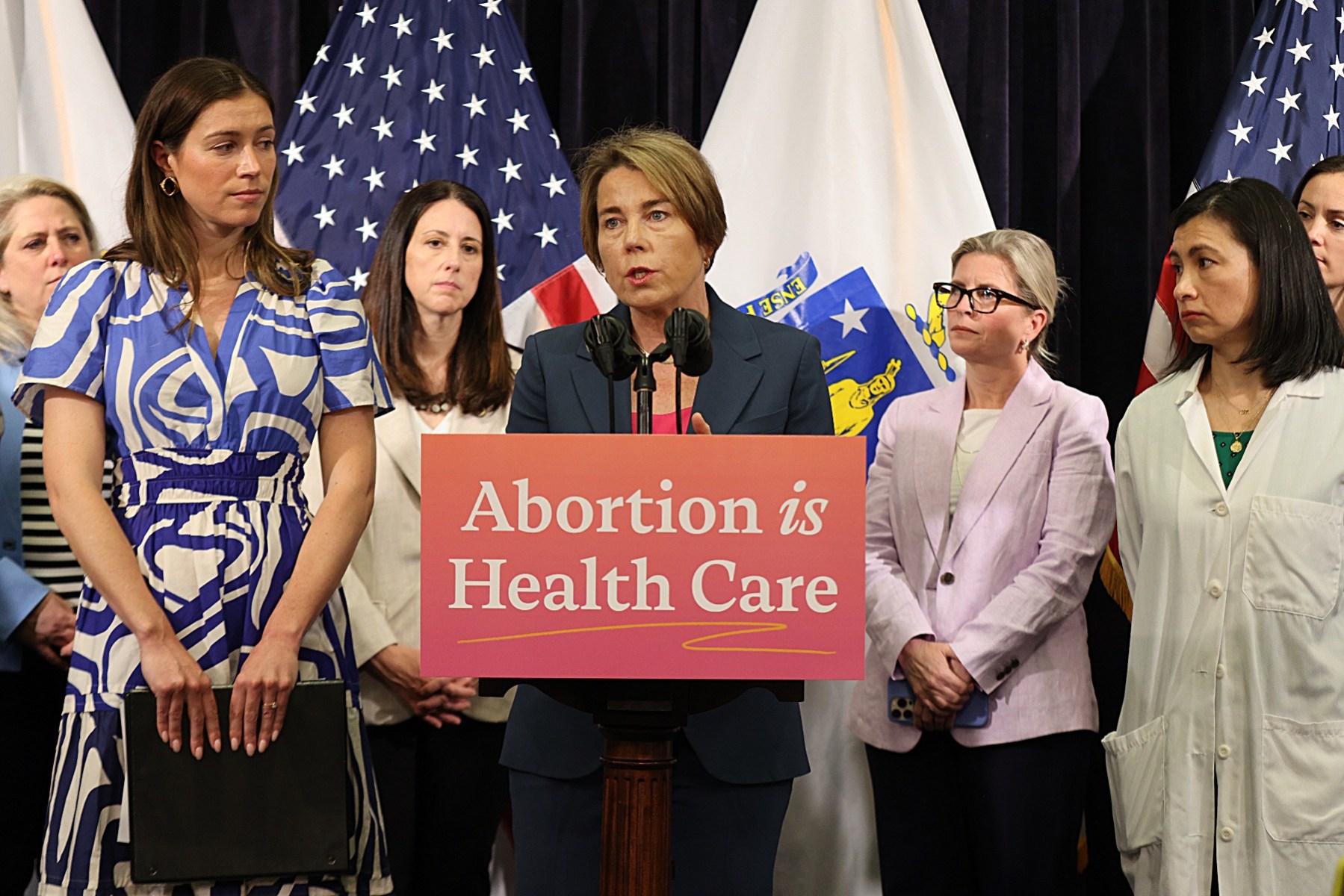 Massachusetts Gov. Maura Healy stands at a podium with "Abortion is Health Care" written on the front as people stand around her.