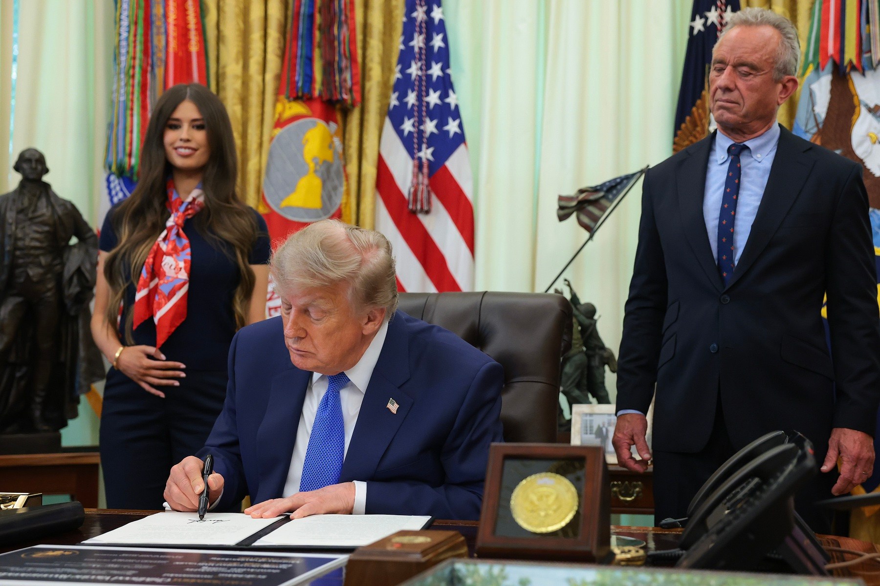 President Donald Trump signs a document with a black pen, with a woman holding her pregnant belly to the left of him and Robert F. Kennedy Jr. to the right.