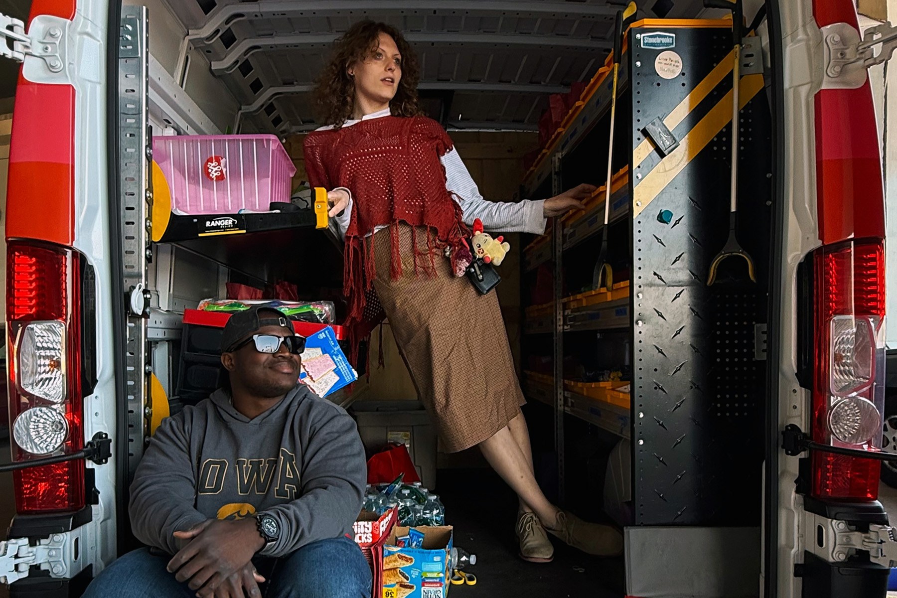 Two outreach workers pose in the open back of a red mobile health van filled with supplies and equipment.