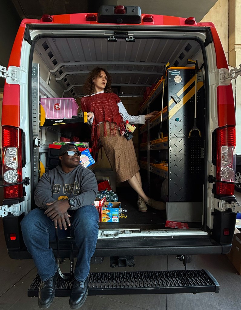 Full-length view of two outreach workers posing in the open back of a red mobile health van, surrounded by storage bins, tools and boxes of supplies.