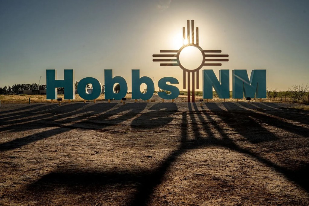 A large roadside “Hobbs NM” sign is silhouetted by the low sun, casting long shadows across the ground.