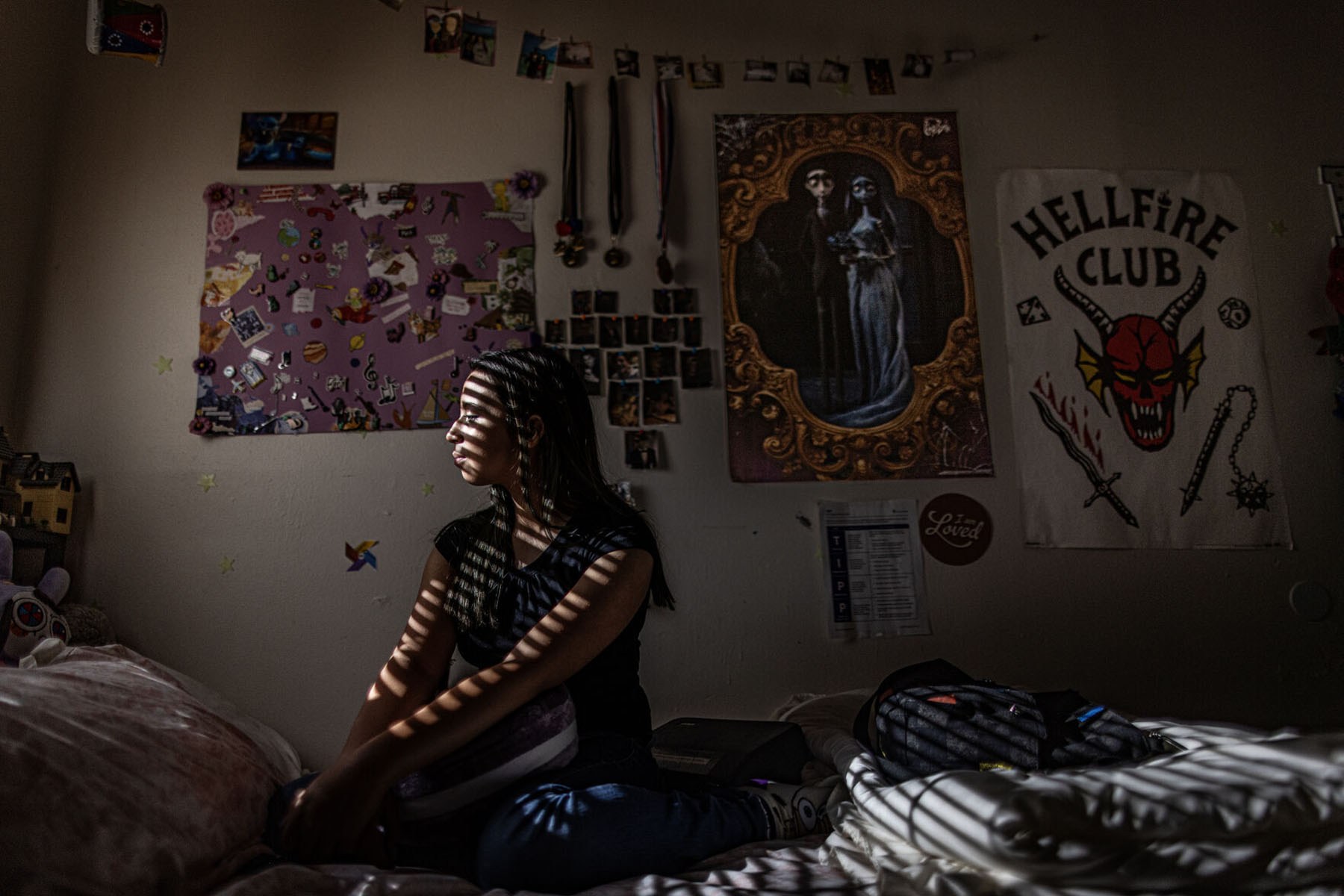 A young girl sits on a bed in a dark bedroom as narrow bands of sunlight from window blinds fall across her face and body.
