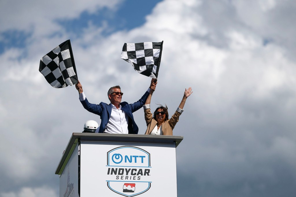 Sean Duffy and Rachel Campos-Duffy stand on an IndyCar platform waving checkered flags beneath a cloudy sky.
