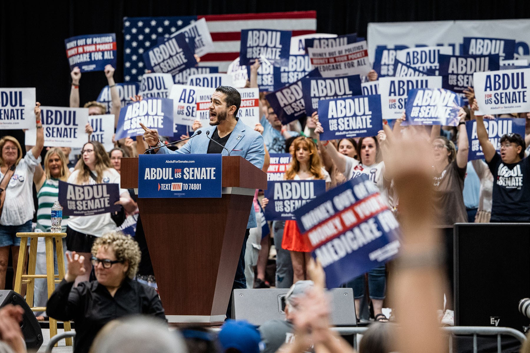 Senate candidate Abdul El-Sayed smiles at a podium as supporters behind him raise campaign signs.