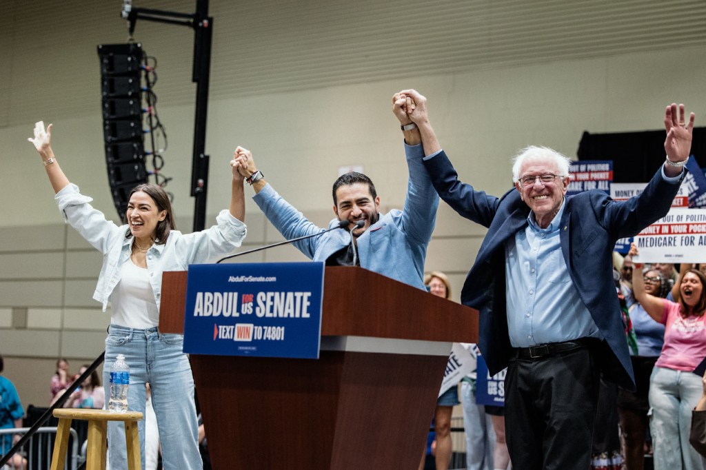 From left, Rep. Alexandria Ocasio-Cortez, Abdul El-Sayed and Sen. Bernie Sanders raise their joined hands onstage as supporters cheer behind them.