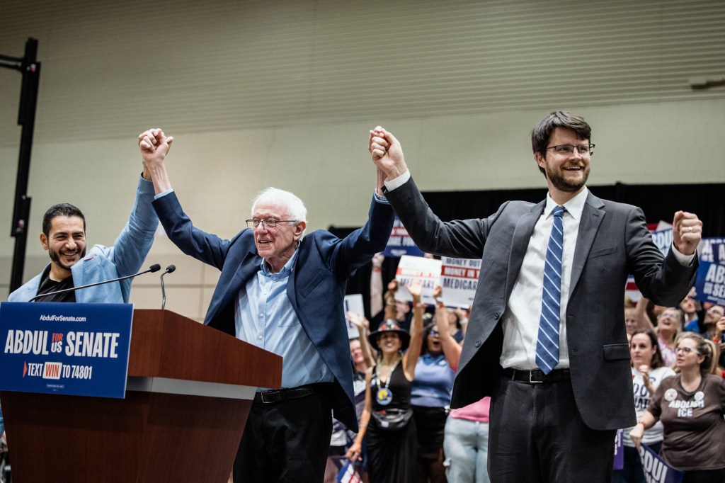 Abdul El-Sayed, Bernie Sanders and Will Lawrence stand onstage with their joined hands raised as supporters cheer behind them.