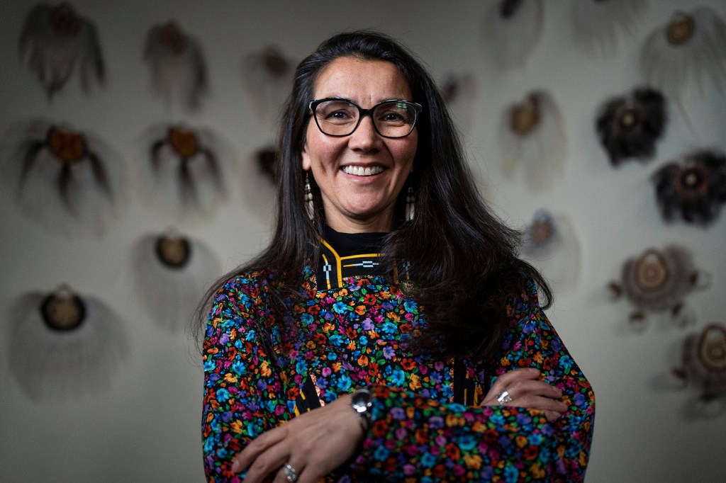 Mary Peltola smiles for a portrait with her arms crossed, wearing glasses and a colorful floral top in front of a wall decorated with woven artwork.