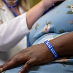 A closeup photo on a woman's wrist wearing the I gave birth wristband, with a nurse checking her vitals in the background.