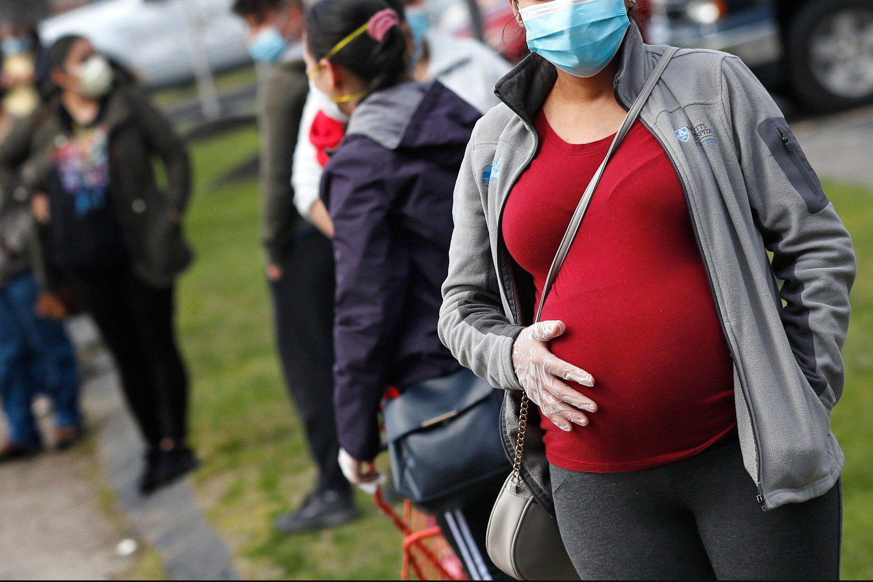 A pregnant woman wearing a mask and gloves stand outside among others in masks.