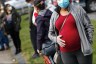 A pregnant woman wearing a mask and gloves stand outside among others in masks.