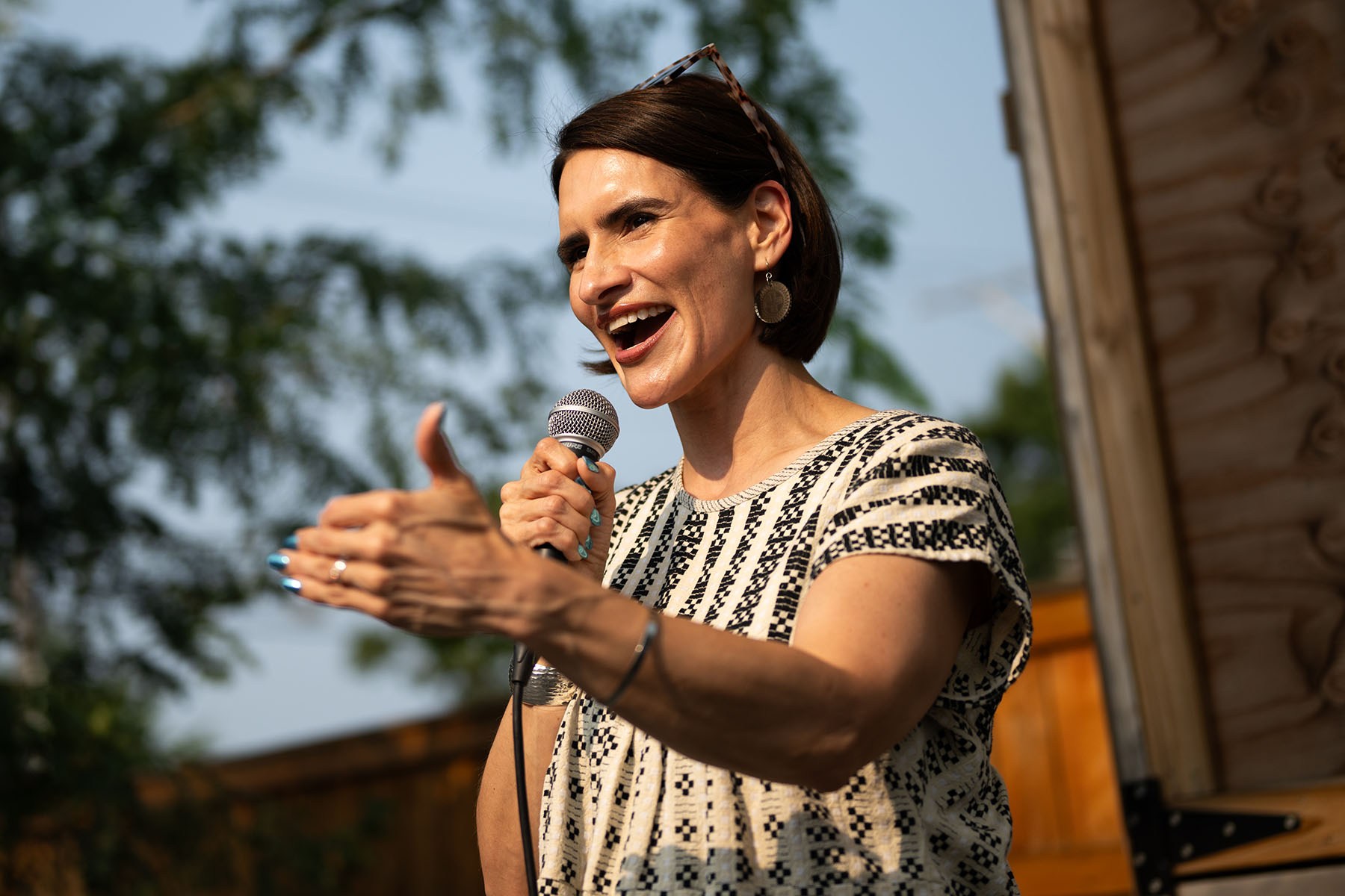 Peggy Flanagan speaks into a microphone at an outdoor event, gesturing with one hand as she addresses the crowd.