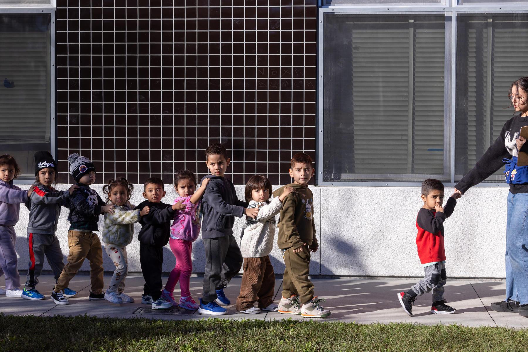 A line of young students hold hands walking outside of a school.