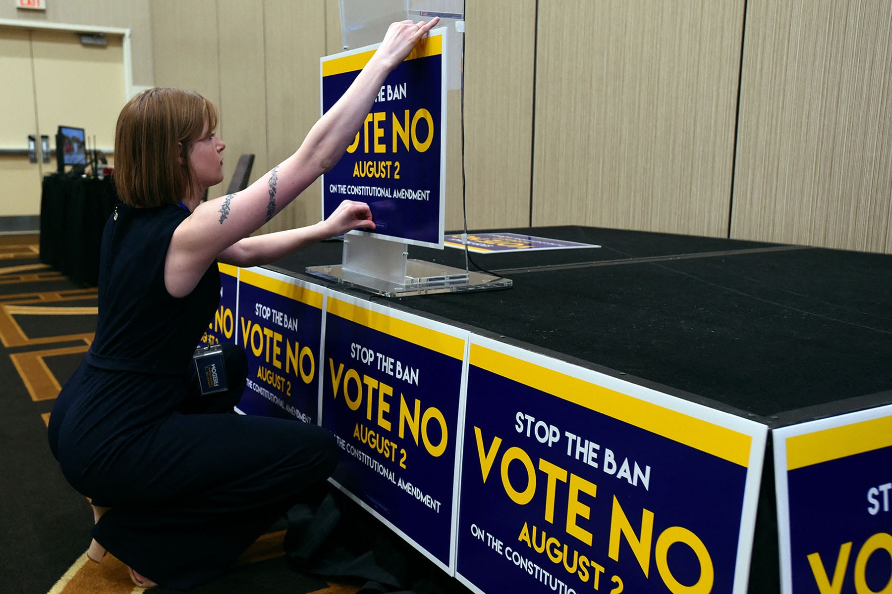 A woman crouches beside a stage, attaching a blue-and-yellow "Vote No" sign to a podium as matching signs cover the front of the stage.