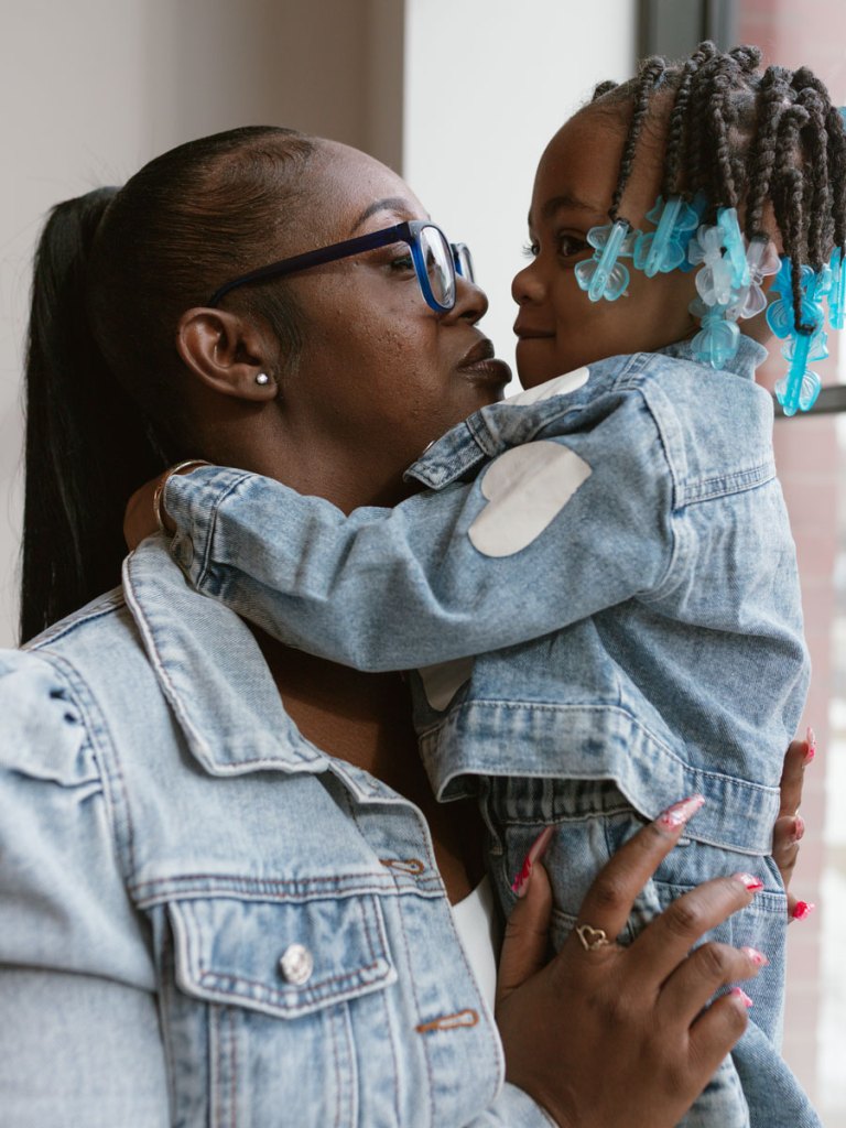 A woman in a denim jacket and glasses holds a young girl with braided hair and blue bow barrettes, the two smiling at each other in an affectionate embrace.