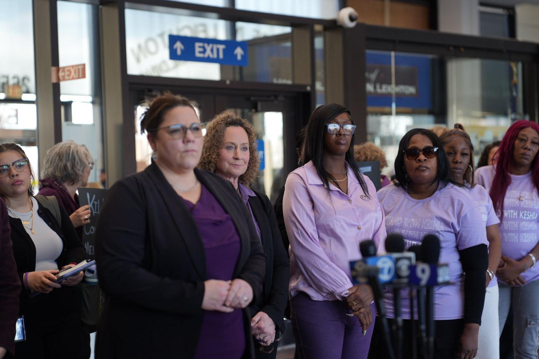A group of women, several wearing purple, including Keshia Golden stand behind microphones during a press conference.