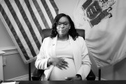 A portrait of McIver in an office surrounded by flags and holding her pregnant belly.