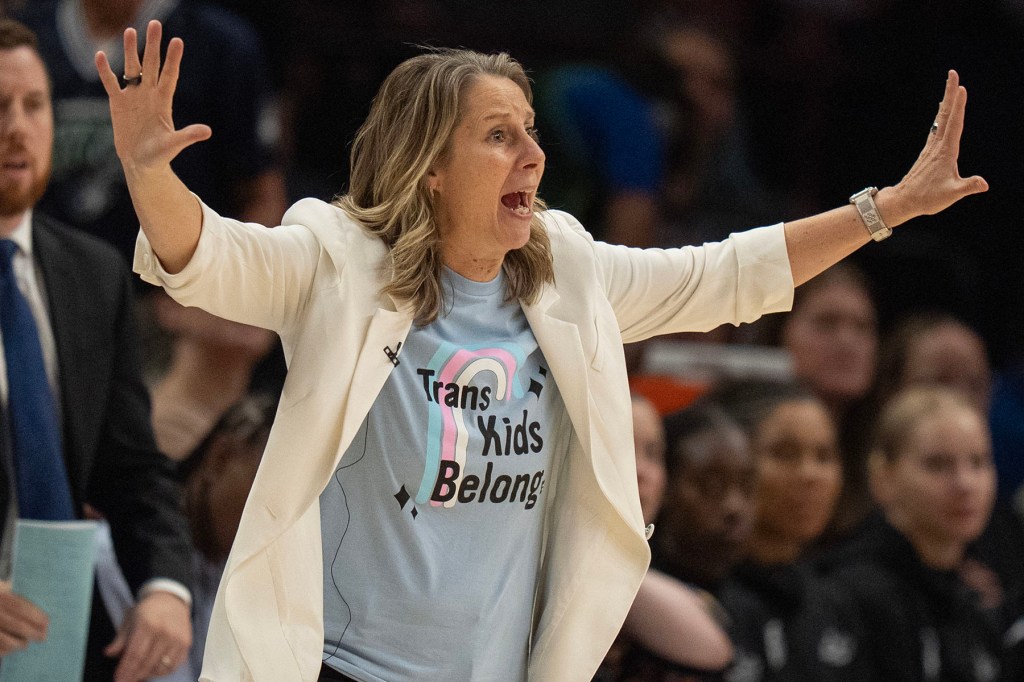 Cheryl Reeve raises both arms while wearing a light blue "Trans Kids Belong" T-shirt beneath a white blazer.