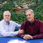 Bob Witeck (left) and Bob Connelly sit together at an outdoor table, one resting a hand on the other's arm, with dense green ivy behind them.