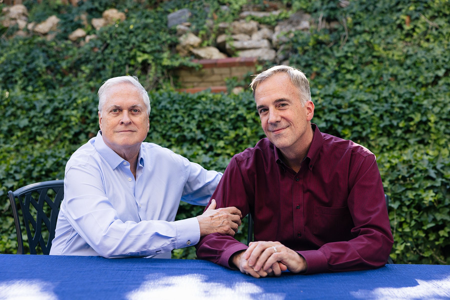 Bob Witeck (left) and Bob Connelly sit together at an outdoor table, one resting a hand on the other's arm, with dense green ivy behind them.