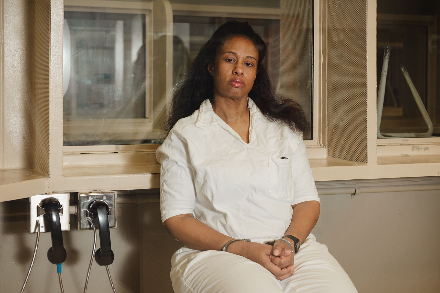 Kwaneta Harris in a white prison uniform sits behind a glass visitation partition with her hands cuffed in her lap. Two black telephone receivers hang on the wall beside her.