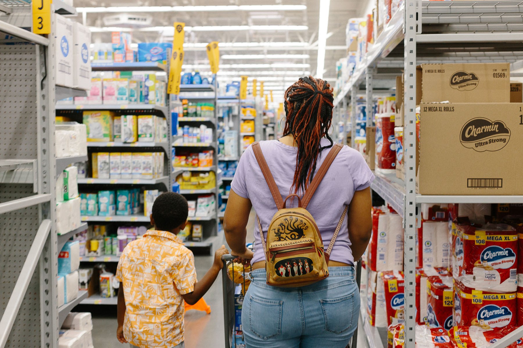 A woman and child walk down a store aisle stocked with household goods, including toilet paper and other everyday essentials.