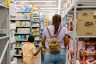 A woman and child walk down a store aisle stocked with household goods, including toilet paper and other everyday essentials.