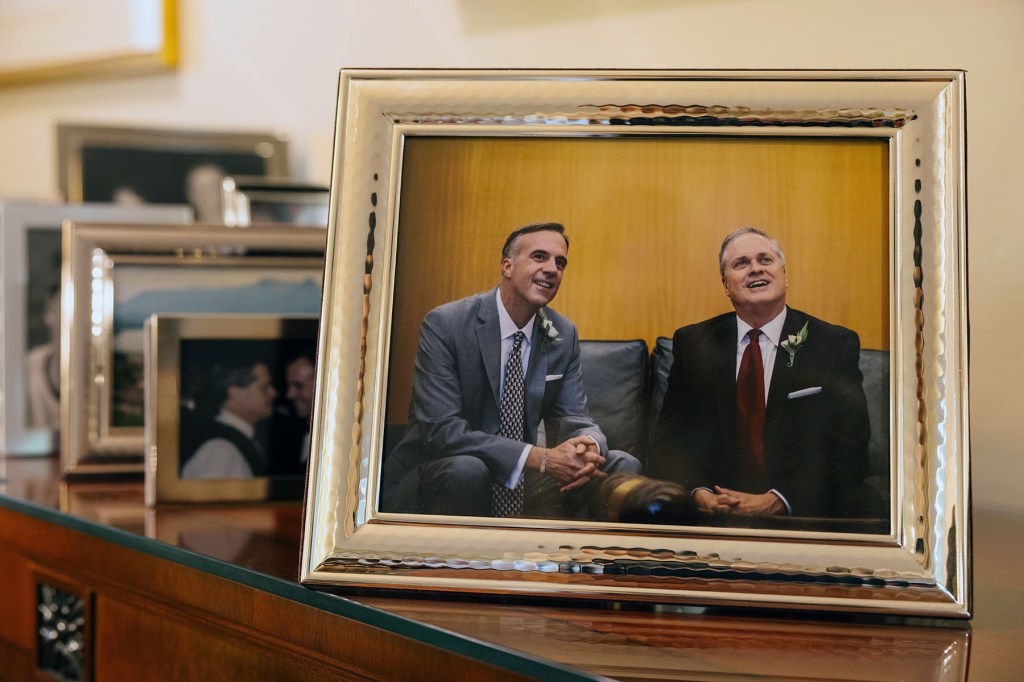 A framed wedding photo of two men in suits sits on a table among other framed photographs.