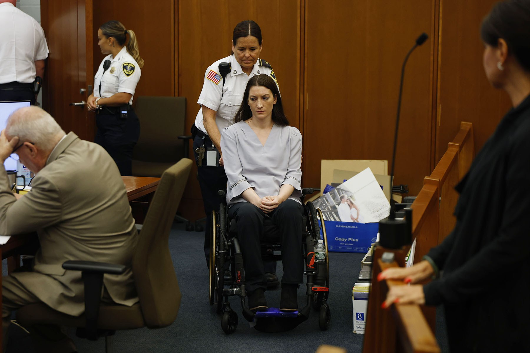 Lindsay Clancy is rolled into a courtroom in a wheelchair with her hands clasped in her lap as a uniformed officer stands behind her. Other court personnel and attorneys are visible around the room.