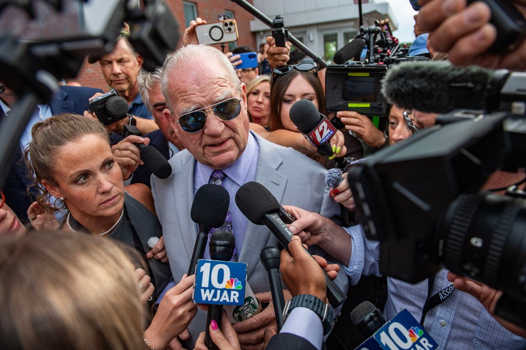 An older man in sunglasses and a light gray suit is surrounded by reporters, cameras and microphones as he speaks outside a building.