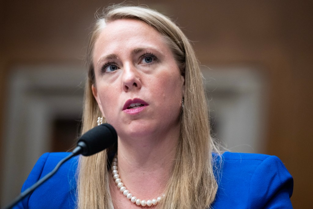 A white woman with blonde hair in a blue jacket speaks into a mic in closeup.
