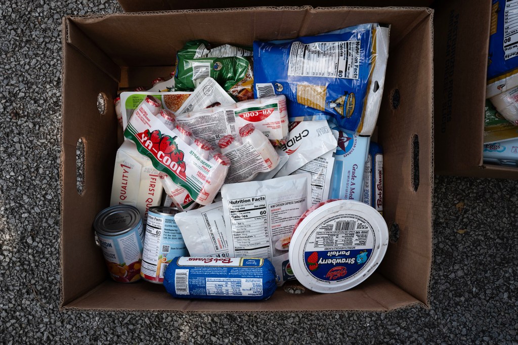 A cardboard box filled with packaged food, including cheese, yogurt, canned goods and other groceries, sits on pavement.