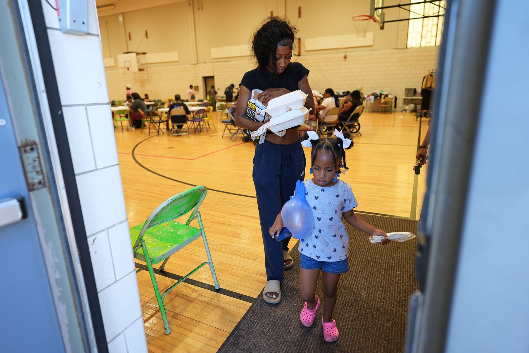 A woman carrying several takeout containers walks out of a gym with a young girl. People sit at tables in the background.