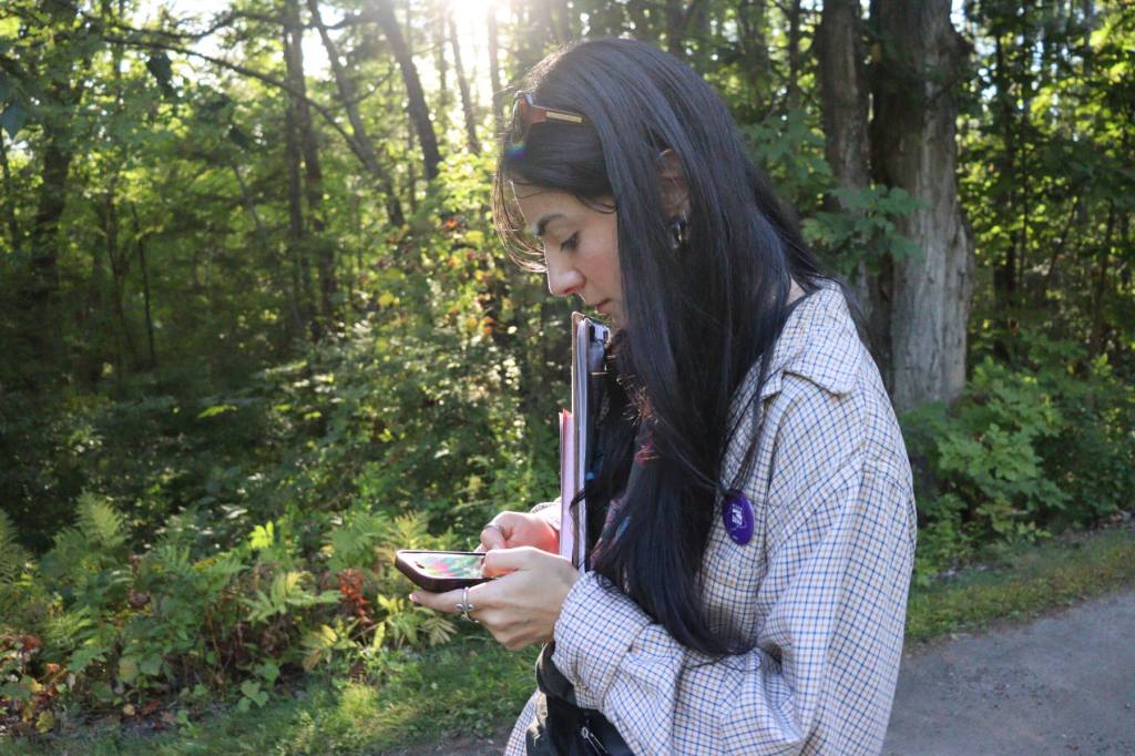 A woman looks at her phone while walking down a street with trees in the backdrop.