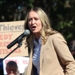 Clayton speaks at a podium with a sign and another woman behind her.