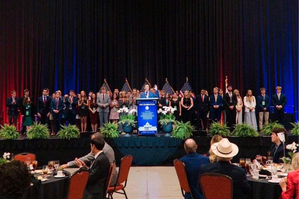 Dozens of young people stand on a stage at a political banquet with others watching from tables situated below.