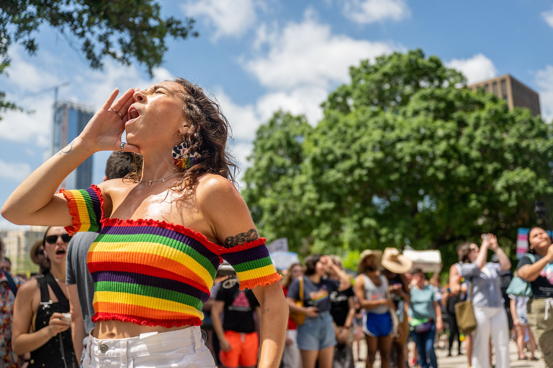 A person in a rainbow-striped off-the-shoulder top cups their hand around their mouth while shouting at a gathering. A crowd stands behind them beneath trees and a bright blue sky.