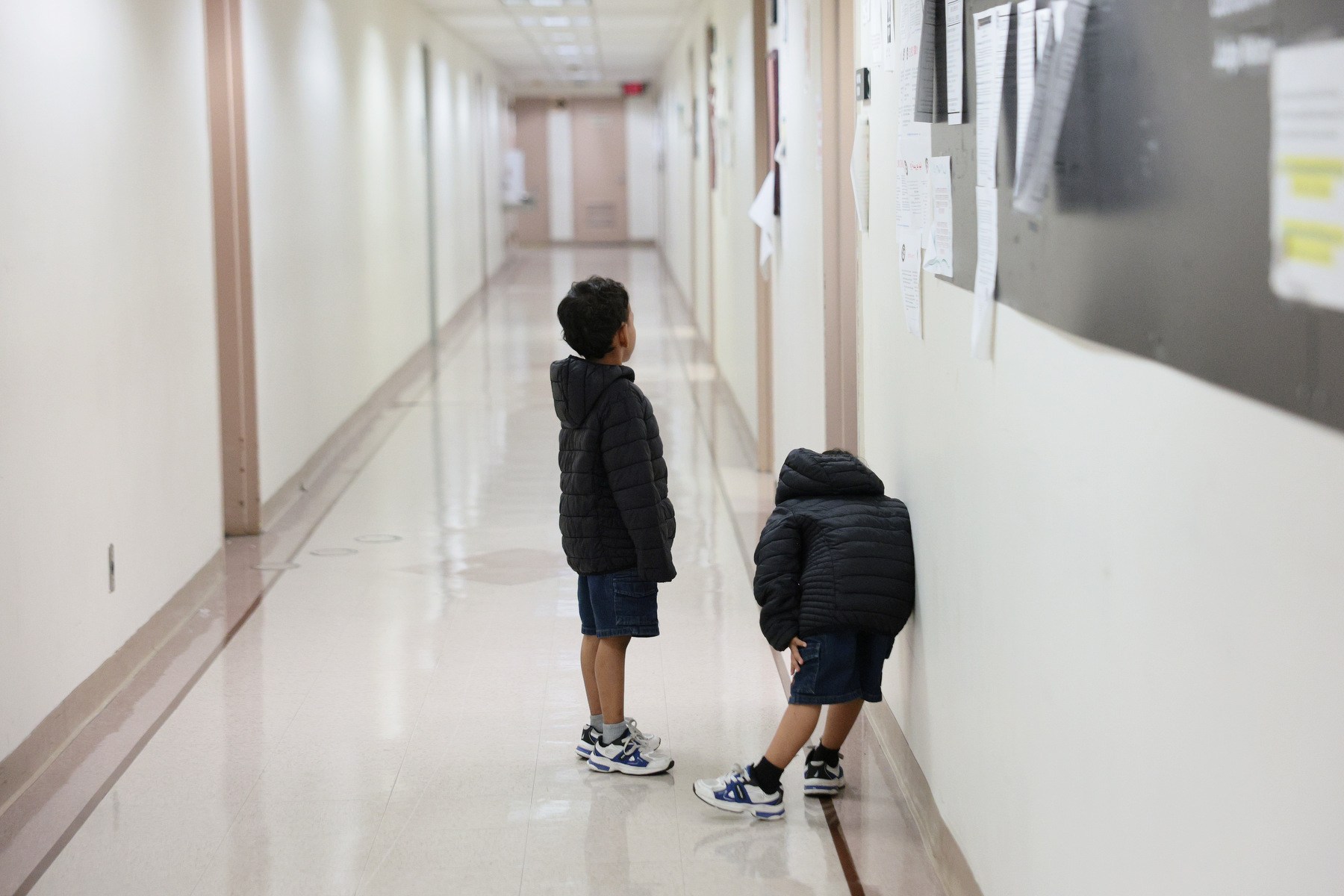 Two children wearing black clothes and facing away from the camera wait in a long hallway alone.