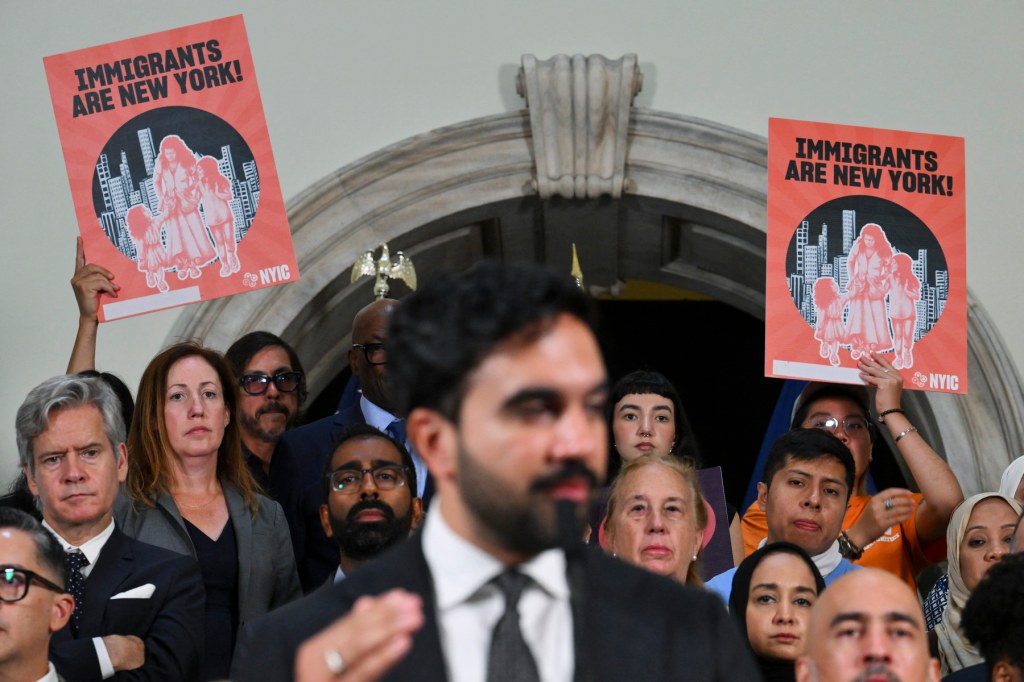 Mamdani speaks at a podium with a crowd of people holding signs standing behind him.