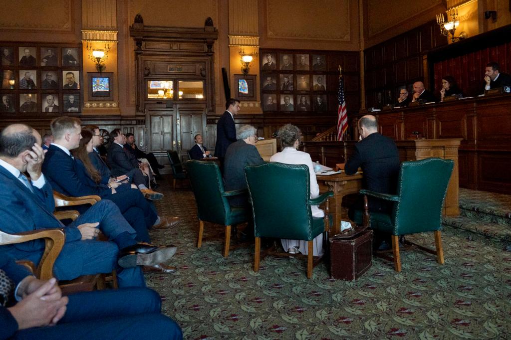 A group of people sit in a courtroom, watching the proceedings as a lawyer stands at a podium and judges watch from above.