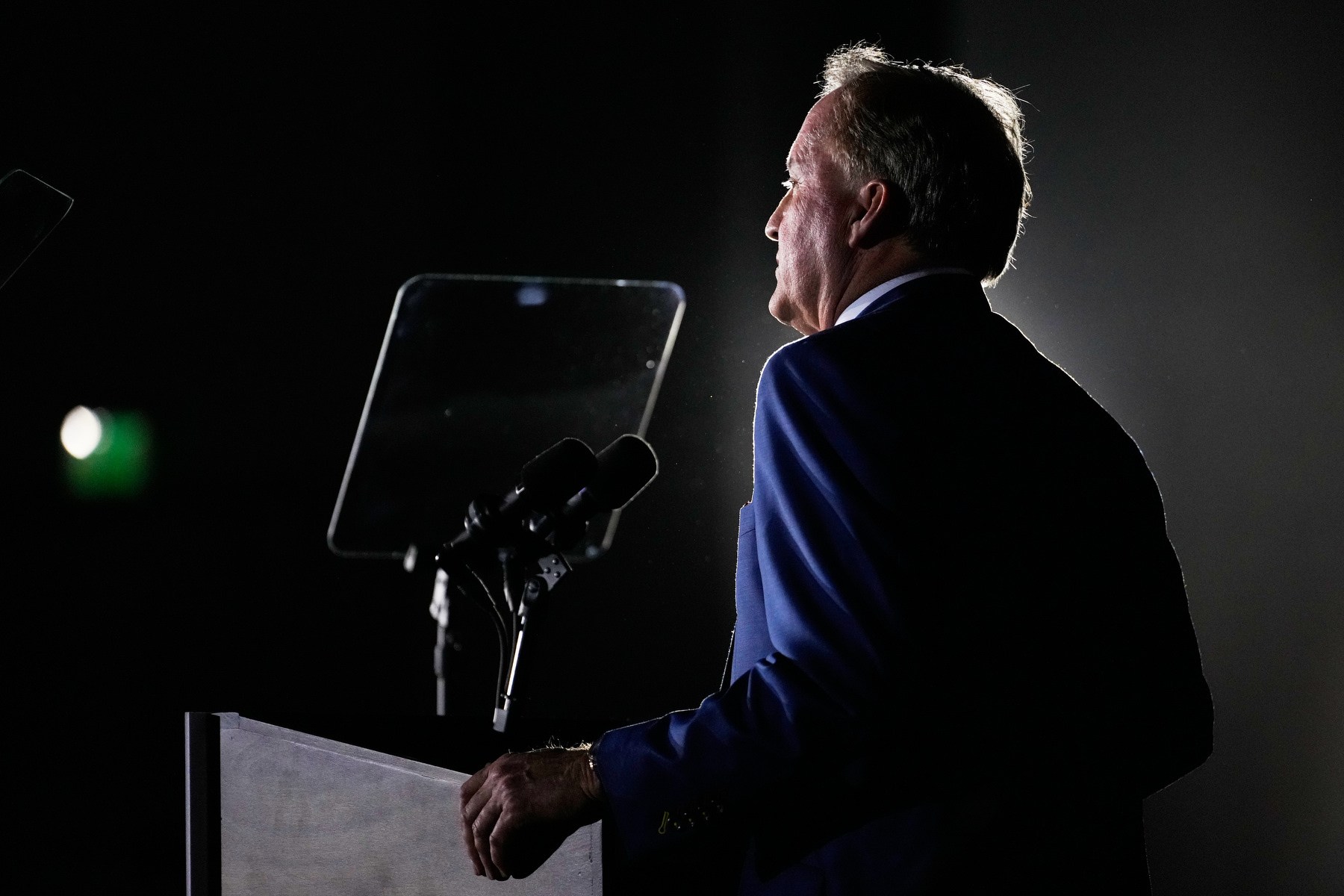 Paxton stands at a podium from behind and backlit.