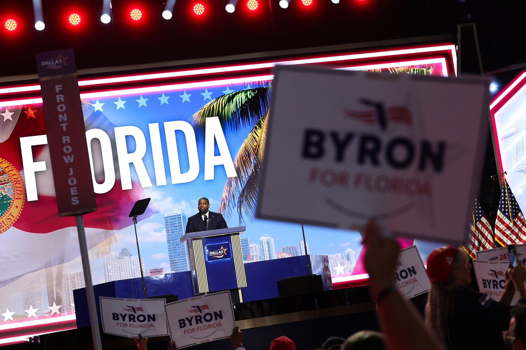 Florida Republican gubernatorial candidate Rep. Byron Donalds stands at a podium in front of a large "Florida" backdrop as audience members hold up "Byron for Florida" signs in the foreground.